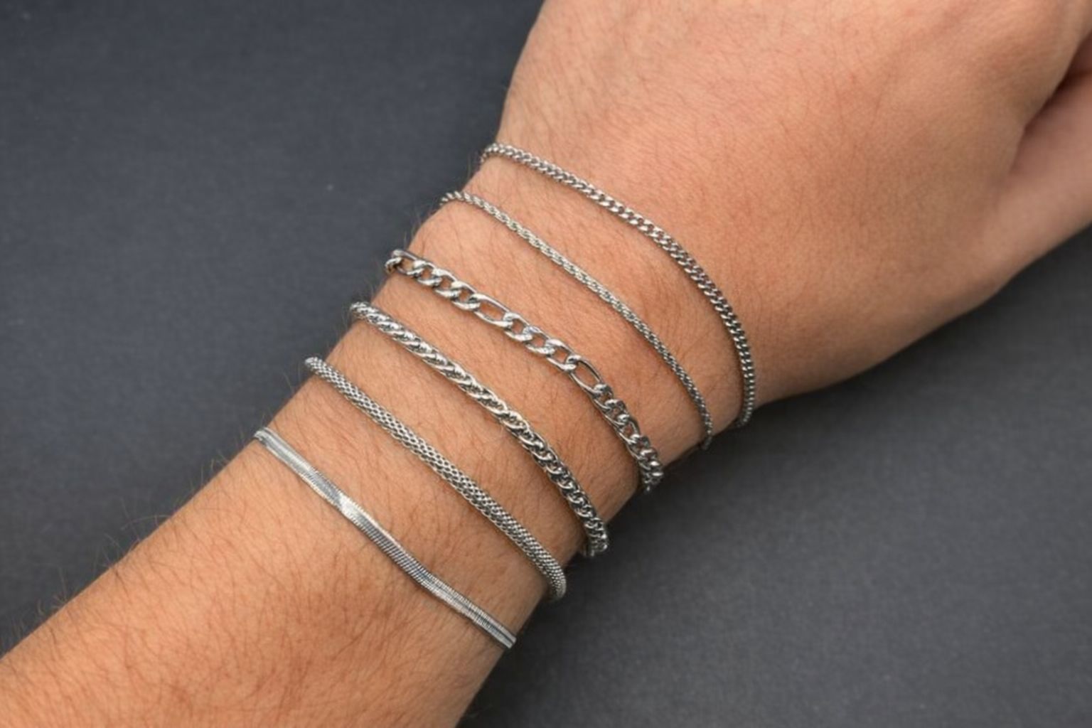 Sju silverfärgade armband av olika design på en arm mot mörk bakgrund.